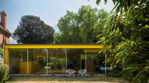 A single storey building with full floor to ceiling windows. The building has a vibrant yellow fascia. A large tree sit in the foreground to the right of the buisling. facia